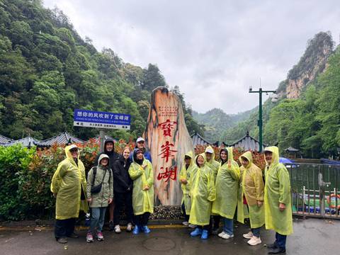       Group wearing ponchos poses beside a carved stone sign and lake within a lush valley park.
  