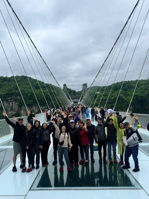       Tourists cheer on a long glass suspension bridge spanning a deep gorge with lush cliffs.
  