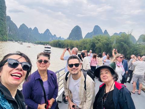       Travelers on a river cruise deck with karst mountains and bamboo groves lining the waterway.
  