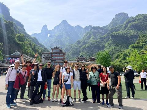       Group photo in lush valley framed by steep green peaks and traditional styled buildings.
  