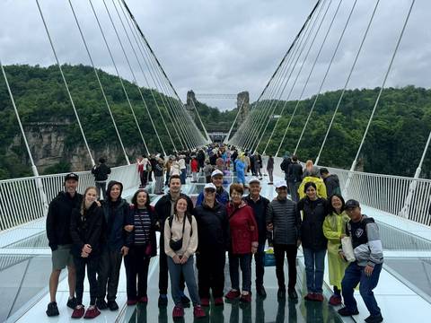       Group lined up for a photo on the glass suspension bridge spanning a gorge with dense greenery.
  