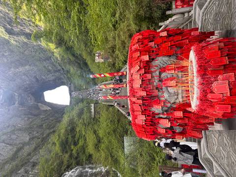       Long stone staircase lined with red prayer plaques and incense leads up to a dramatic natural arch set in lush green cliffs.
  