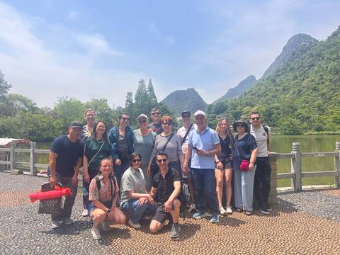       Smiling tour group poses beside a tranquil lake with jagged karst peaks and greenery under a blue sky.
  