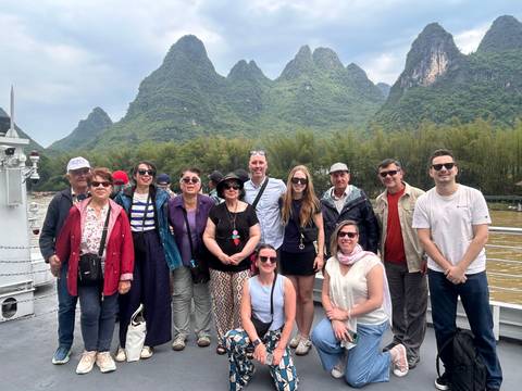       Tourists stand on a riverboat deck with lush karst mountains rising behind them.
  