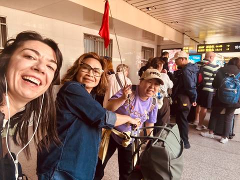       Three travelers smiling and making peace signs while waiting on a busy train platform with luggage.
  