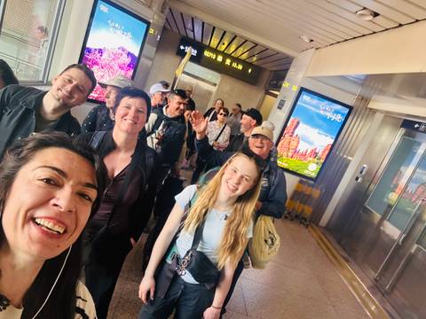       Cheerful group selfie inside a modern rail station corridor with digital signs overhead.
  