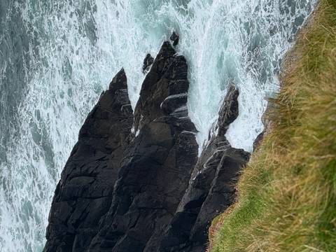       Wild Atlantic waves crash against dark jagged rocks below a grassy cliff edge.
  