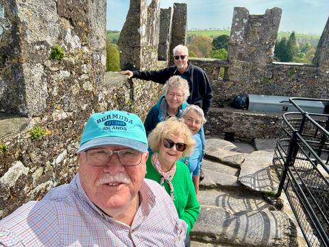       Older travelers smile on stone steps within a weathered castle ruin surrounded by greenery.
  