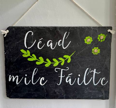       Slate welcome sign painted in Gaelic with green leaf design hanging on a wall.
  