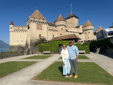       Couple posing in front of medieval lakeside Château de Chillon with clear blue sky.
  