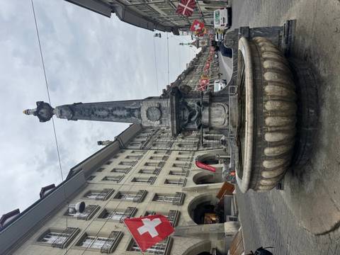       Historic stone fountain with obelisk center on a cobbled street lined by classic buildings and flags.
  