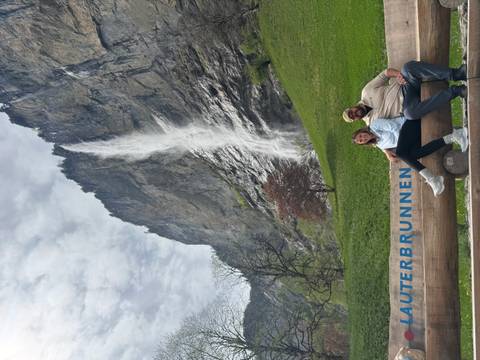       Couple sitting on a log with a tall waterfall and cliff face towering behind them in Lauterbrunnen.
  