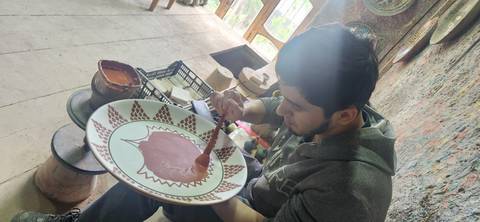       Young artisan painting intricate patterns on a large ceramic plate inside a workshop.
  