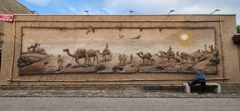       Large relief mural of camel caravans on a wall with a seated man resting on a bench.
  
