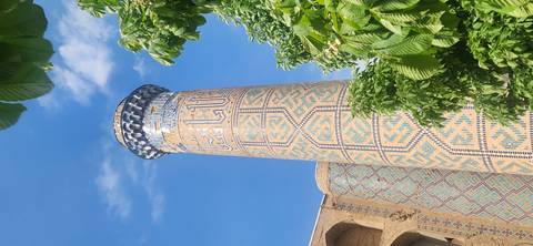       Tall slender minaret covered in intricate turquoise and yellow tilework rises toward a blue sky framed by greenery.
  