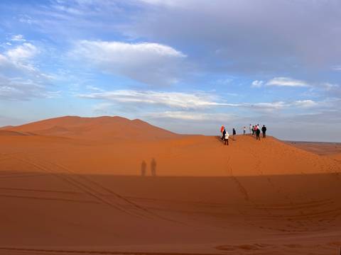      Travellers walk along the crest of vast orange sand dunes at golden hour casting long shadows across the Sahara.
  