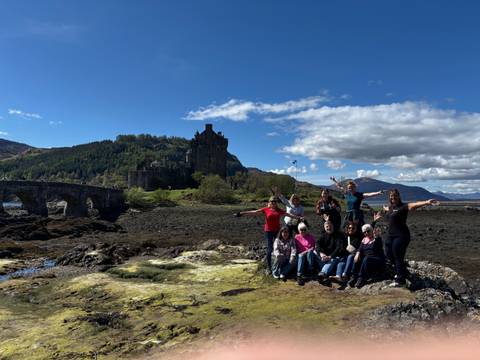       Small tour group poses happily in front of Eilean Donan Castle with mountains behind.
  