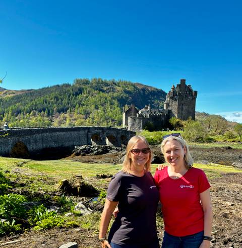       Two smiling women stand before a stone bridge and castle against a green hillside.
  