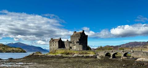       Historic Eilean Donan Castle sits on a rocky islet with surrounding mountains and bridge under blue skies.
  