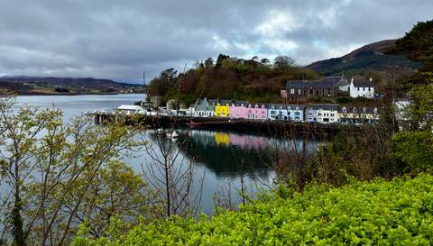       Colourful waterfront houses of Portree reflect in the calm harbour surrounded by green hills.
  