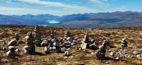       Rock cairns dot a windswept moorland plateau overlooking distant lochs and mountain ranges.
  