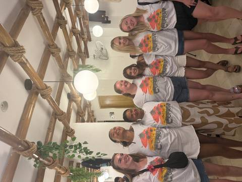       Group of travellers wearing matching tour shirts pose indoors under bamboo ceiling decor.
  