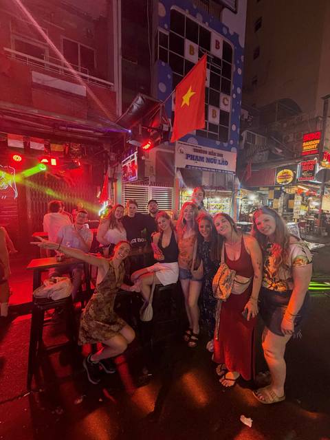       A lively group of backpackers poses inside a neon-lit bar street at night.
  