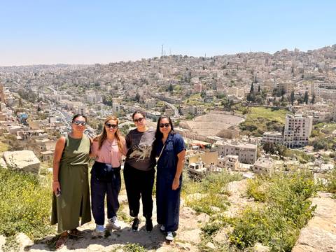       Four women smile with the Roman Theater of Amman and sprawling cityscape behind them.
  
