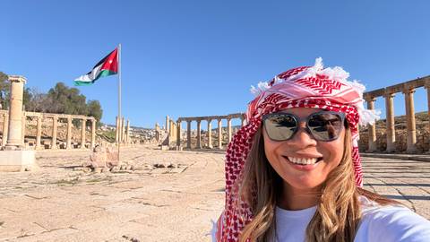       Selfie of a smiling woman wearing a red-and-white keffiyeh with Jerash’s oval colonnade and Jordan flag behind her.
  