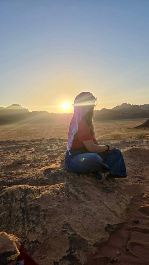       Silhouetted woman wearing a keffiyeh gazes at a desert sunset in Wadi Rum.
  