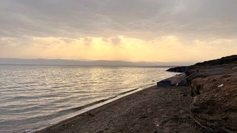       Calm Dead Sea shoreline at dusk with golden rays breaking through clouds across the water.
  