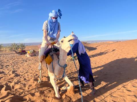       Traveller in blue headscarf rides a white camel led by a local guide across orange desert dunes.
  
