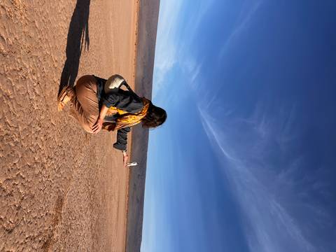       Traveller squats on cracked desert ground creating a forced-perspective pose with a distant friend under vivid blue sky.
  