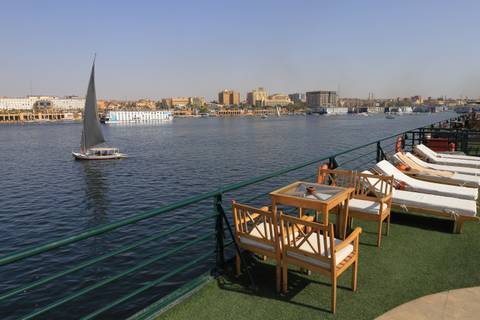       Sun loungers and tables line the deck of a Nile cruise boat overlooking calm river waters and a passing felucca sailboat.
  