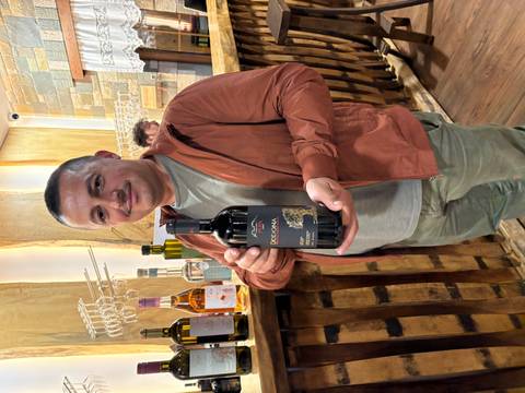       A smiling man in a winery proudly presents a bottle of Albanian red wine.
  