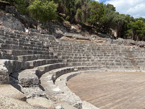       Ancient stone amphitheatre tiers curving around a wooden stage with one visitor walking along the top row
  