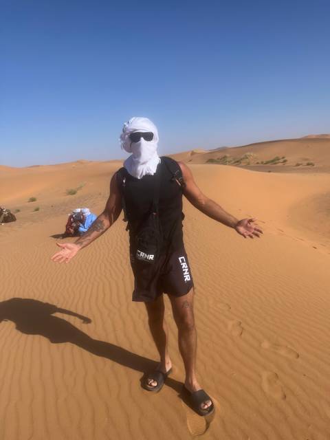       Traveler wearing headscarf and sunglasses stands amid rolling Sahara dunes under harsh sun.
  