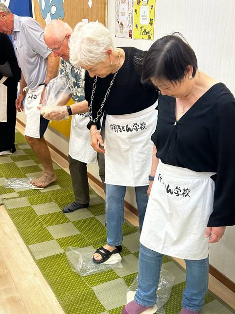       Participants wearing aprons stand barefoot or in socks on tatami mats during a Japanese noodle-making class.
  