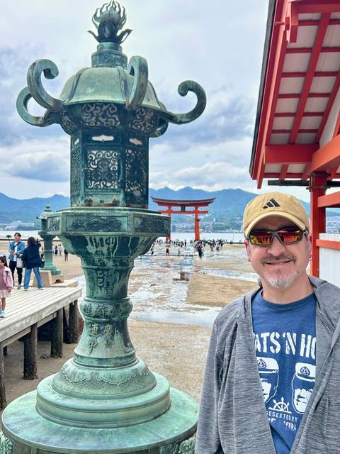       Smiling tourist in front of bronze lantern with the famous floating Torii gate of Miyajima Island and distant mountains.
  