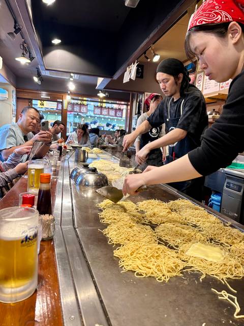       Chefs prepare heaps of noodles on a large teppan grill while diners sit at the counter in a bustling Japanese eatery.
  