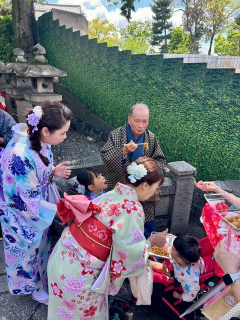       Family in colorful yukata tasting street food beside a stone fence and lush green wall.
  
