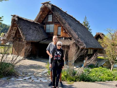       Older couple poses in front of a traditional gassho-zukuri thatched farmhouse in Shirakawa-go on a sunny morning.
  