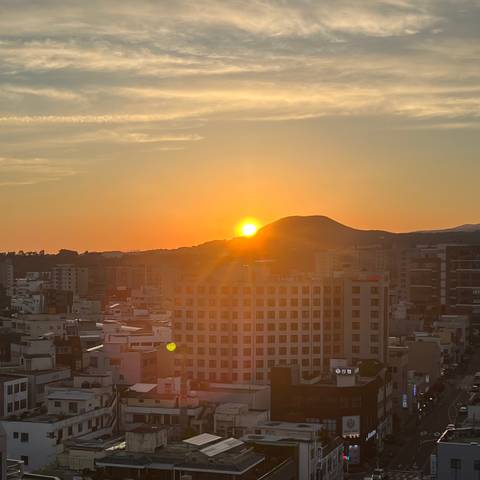       Orange sun setting behind a small hill above a cityscape of mid-rise buildings.
  