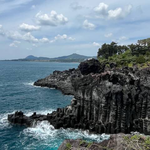      Dramatic basalt column cliffs meeting a deep blue ocean with distant mountains under a partly cloudy sky.
  