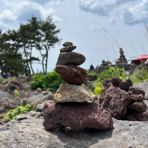       Small balanced stack of volcanic rocks with blurred natural background of shrubs and distant cairns.
  