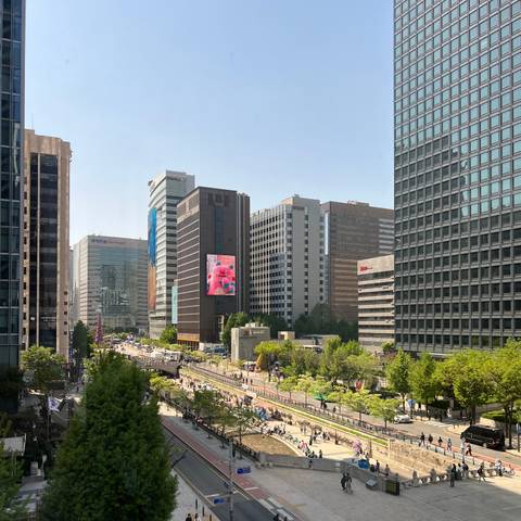       Modern downtown skyline with tall glass buildings and a digital billboard above a tree-lined stream.
  