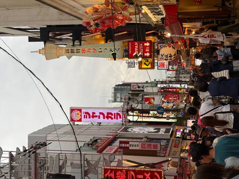       Bustling pedestrian street lined with neon signs and shopfronts under an overcast evening sky.
  