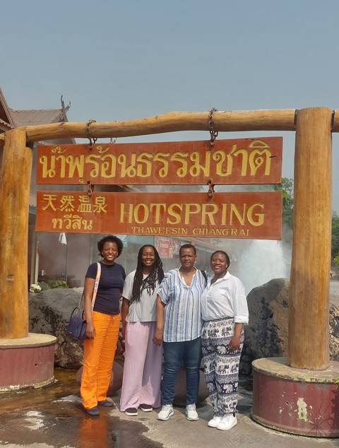      Four women posing beside a wooden signboard for a natural hot spring attraction.
  