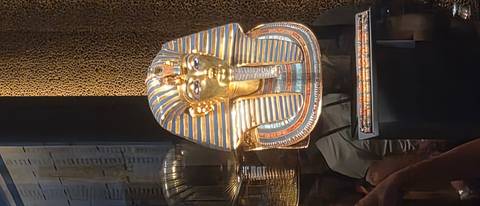       Golden funerary mask of Tutankhamun displayed behind museum glass under spotlights.
  