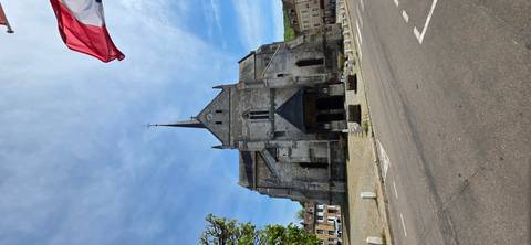       Stone church with tall spire in small town square beneath blue sky and flag.
  